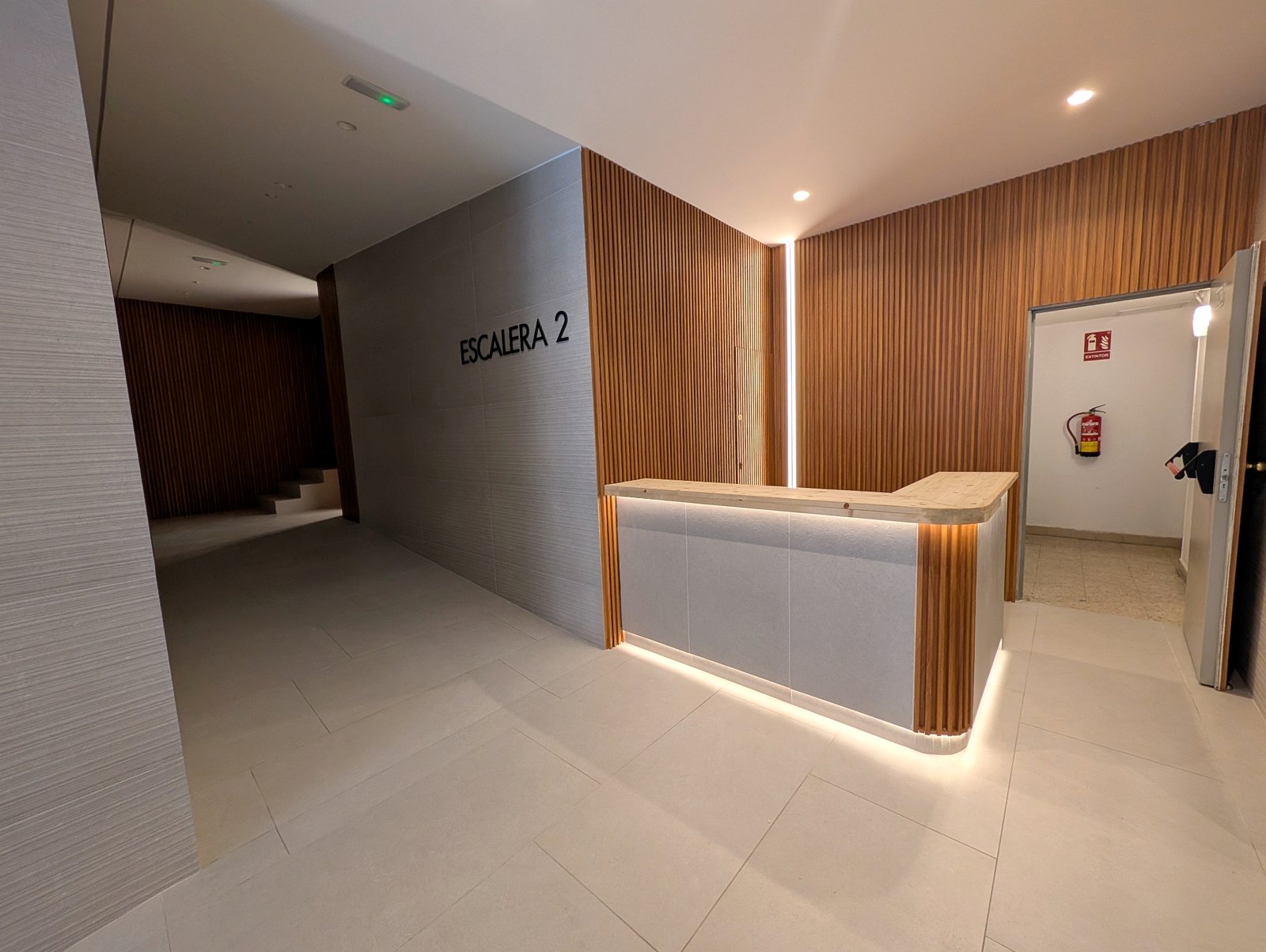 Portal rehabilitado en Madrid con panel de lamas de madera, iluminación LED integrada, señalética moderna y mostrador de conserjería a medida - Grupo Illescas